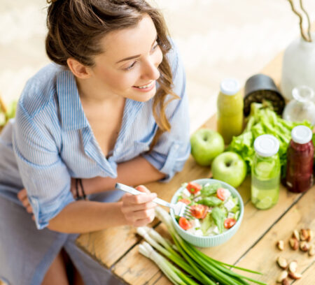 Young and happy woman eating healthy salad sitting on the table with green fresh ingredients indoors