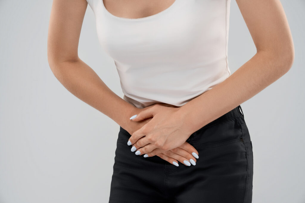 Close up of bending incognito caucasian woman in casual suit posing over gray isolated background and touching lower abdomen because of ache. Front crop of brunette suffering from pain in stomach.